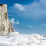 The iconic chalk cliffs of the Seven Sisters rise starkly from the Sussex coastline, their gleaming white faces contrasting against a deep blue sky and the churning waves below. Towering, sheer, and weather-beaten by centuries of sea spray, the cliffs create one of England’s most dramatic coastal views, where walkers trace the narrow paths above and the restless Channel pounds the shingle shore beneath