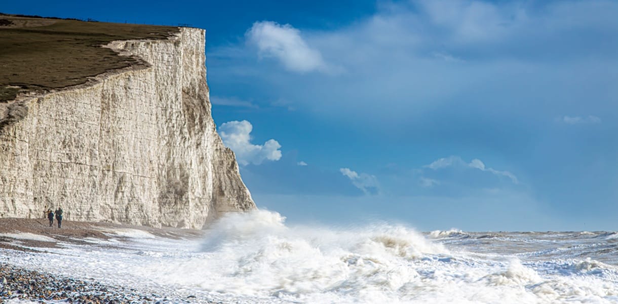 The iconic chalk cliffs of the Seven Sisters rise starkly from the Sussex coastline, their gleaming white faces contrasting against a deep blue sky and the churning waves below. Towering, sheer, and weather-beaten by centuries of sea spray, the cliffs create one of England’s most dramatic coastal views, where walkers trace the narrow paths above and the restless Channel pounds the shingle shore beneath