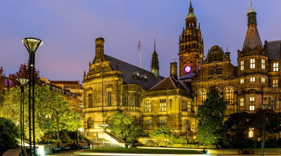 Sheffield City Hall illuminated at dusk, with warm golden lighting highlighting the grand stone façade and clock tower, surrounded by trees and modern lampposts in the Peace Gardens