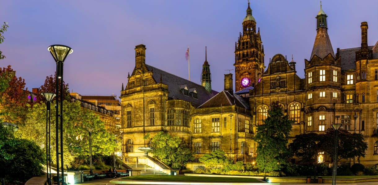 Sheffield City Hall illuminated at dusk, with warm golden lighting highlighting the grand stone façade and clock tower, surrounded by trees and modern lampposts in the Peace Gardens