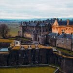 Aerial view of Stirling Castle in Scotland, perched dramatically on a rocky hilltop with sweeping views of the surrounding countryside. The fortress showcases its mix of medieval stone walls and Renaissance-style buildings, with one section lit in a warm golden hue that contrasts with the grey stone. Rolling fields stretch into the distance, framed by distant mountains under a cloudy sky