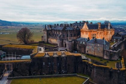 Aerial view of Stirling Castle in Scotland, perched dramatically on a rocky hilltop with sweeping views of the surrounding countryside. The fortress showcases its mix of medieval stone walls and Renaissance-style buildings, with one section lit in a warm golden hue that contrasts with the grey stone. Rolling fields stretch into the distance, framed by distant mountains under a cloudy sky