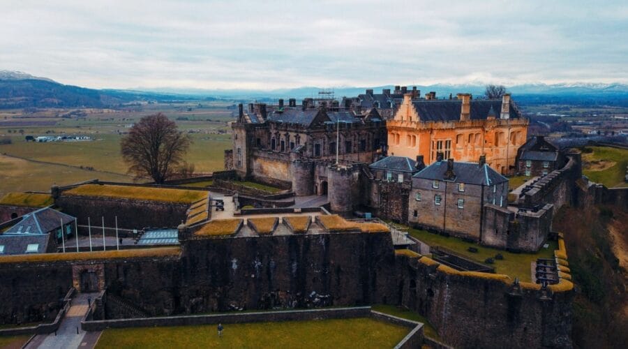 Aerial view of Stirling Castle in Scotland, perched dramatically on a rocky hilltop with sweeping views of the surrounding countryside. The fortress showcases its mix of medieval stone walls and Renaissance-style buildings, with one section lit in a warm golden hue that contrasts with the grey stone. Rolling fields stretch into the distance, framed by distant mountains under a cloudy sky