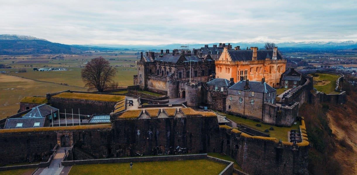 Aerial view of Stirling Castle in Scotland, perched dramatically on a rocky hilltop with sweeping views of the surrounding countryside. The fortress showcases its mix of medieval stone walls and Renaissance-style buildings, with one section lit in a warm golden hue that contrasts with the grey stone. Rolling fields stretch into the distance, framed by distant mountains under a cloudy sky