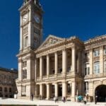 Exterior view of Birmingham Museum and Art Gallery with its grand clock tower and neoclassical façade, featuring tall columns and ornate stonework, under a clear blue sky with people walking and sitting in the square outside.