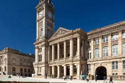 Exterior view of Birmingham Museum and Art Gallery with its grand clock tower and neoclassical façade, featuring tall columns and ornate stonework, under a clear blue sky with people walking and sitting in the square outside.