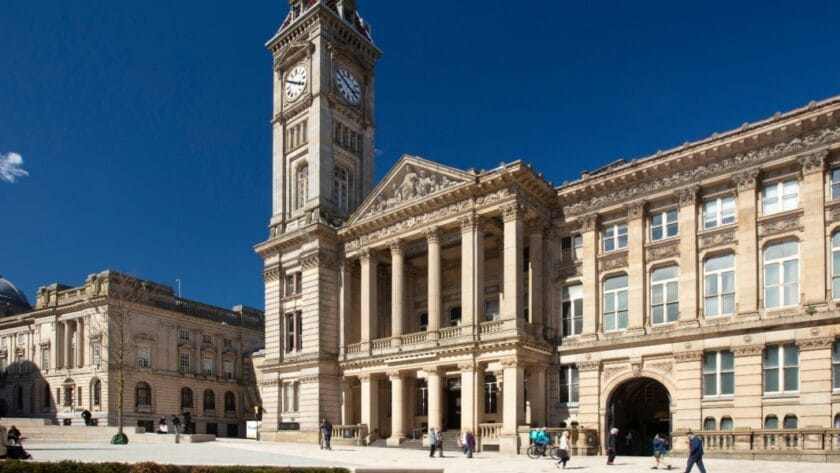 Exterior view of Birmingham Museum and Art Gallery with its grand clock tower and neoclassical façade, featuring tall columns and ornate stonework, under a clear blue sky with people walking and sitting in the square outside.