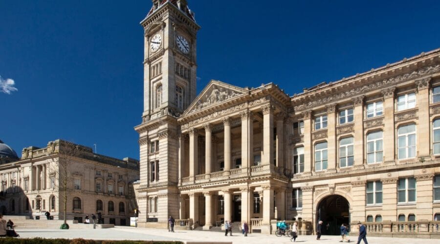 Exterior view of Birmingham Museum and Art Gallery with its grand clock tower and neoclassical façade, featuring tall columns and ornate stonework, under a clear blue sky with people walking and sitting in the square outside.