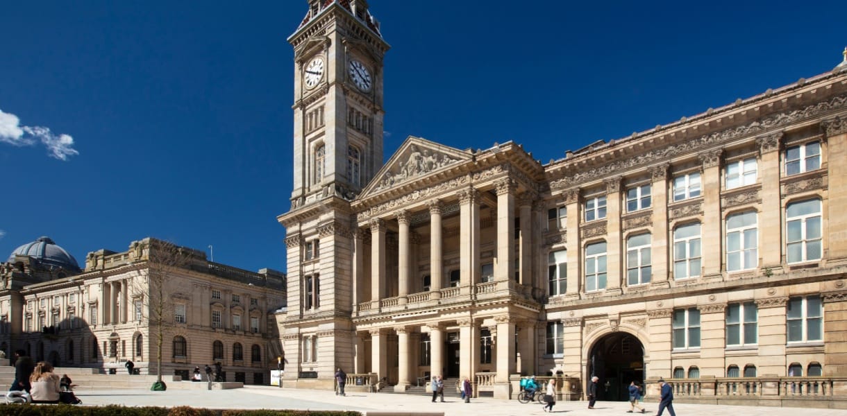 Exterior view of Birmingham Museum and Art Gallery with its grand clock tower and neoclassical façade, featuring tall columns and ornate stonework, under a clear blue sky with people walking and sitting in the square outside.