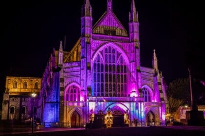 Winchester Cathedral illuminated at night with vibrant purple, pink, and blue lights highlighting its grand Gothic façade, tall arched windows, and intricate stonework against the dark sky