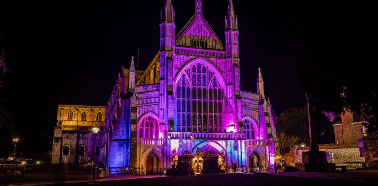 Winchester Cathedral illuminated at night with vibrant purple, pink, and blue lights highlighting its grand Gothic façade, tall arched windows, and intricate stonework against the dark sky