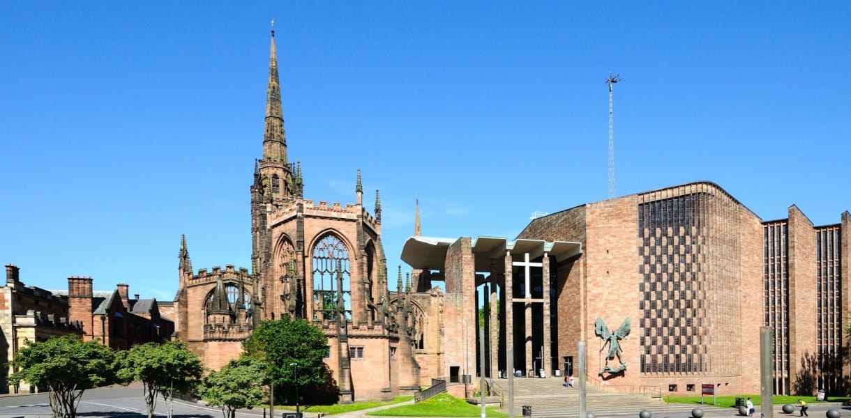 Coventry Cathedral in sunlight, showing the roofless stone ruins of the old Gothic cathedral with its tall spire beside the modern cathedral building, which has a broad brick facade, a large cross and steps leading to the entrance under a clear blue sky