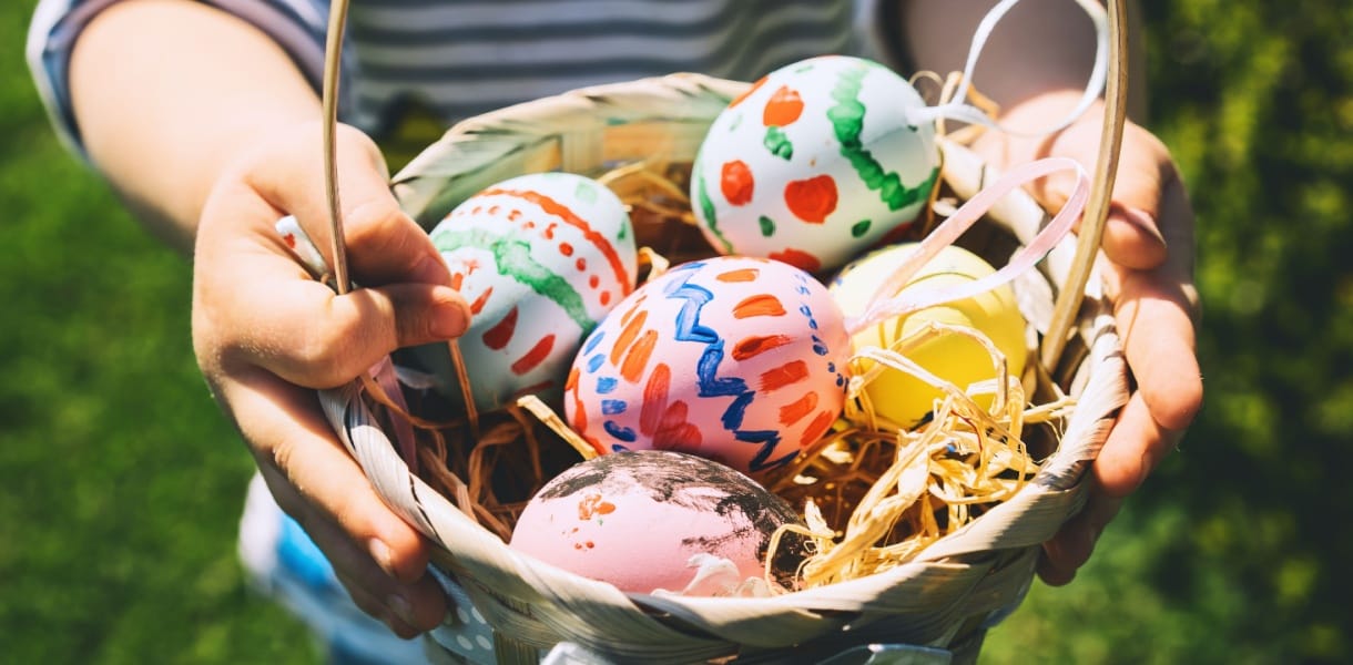 A person outdoors holds a wicker basket filled with straw and hand-painted Easter eggs in pastel colours, including pink, yellow, white and blue, decorated with dots, zigzags and floral patterns