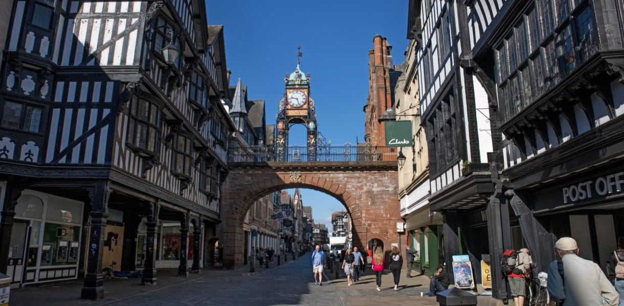 Eastgate Clock in Chester above the historic Eastgate Street, framed by black and white timber framed buildings and a sandstone arch, with people walking below on a bright sunny day.