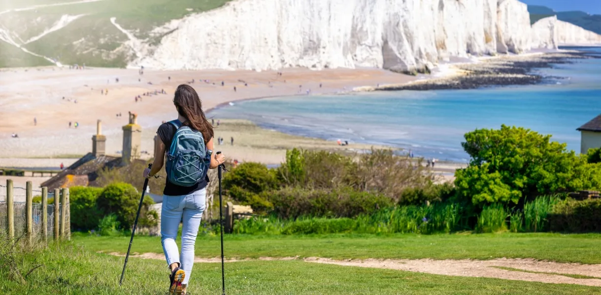 A walker with hiking poles and a backpack looks out over a wide shingle beach and bright blue sea, with towering white chalk cliffs in the background on England’s coast.