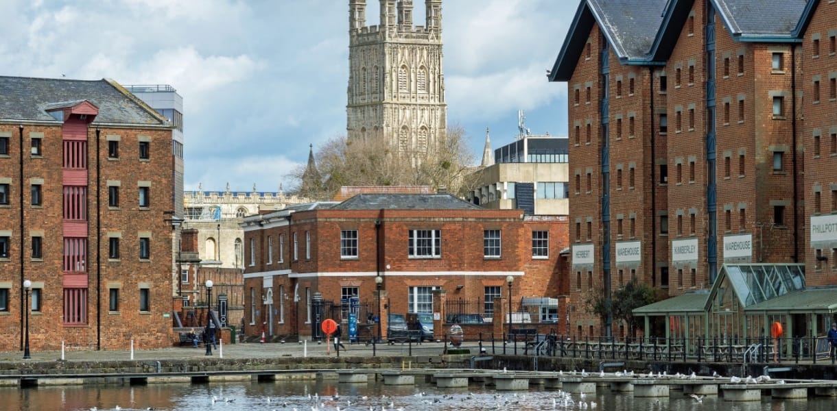 Gloucester Docks with historic red-brick warehouses beside the water and Gloucester Cathedral tower rising in the background under a cloudy sky.
