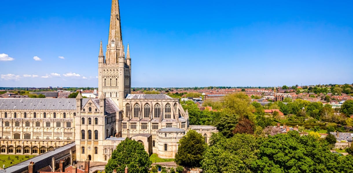 Panoramic view of Norwich Cathedral rising above the city, with its tall stone spire, Romanesque cloisters, and surrounding green trees under a bright blue sky