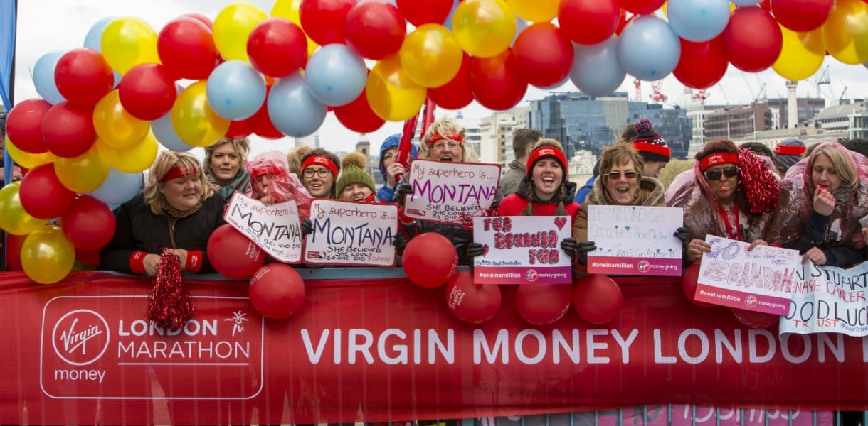 A crowd of cheerful London Marathon supporters stands behind a red Virgin Money barrier and a bright arch of red, yellow, and blue balloons, holding handmade signs and cheering on runners