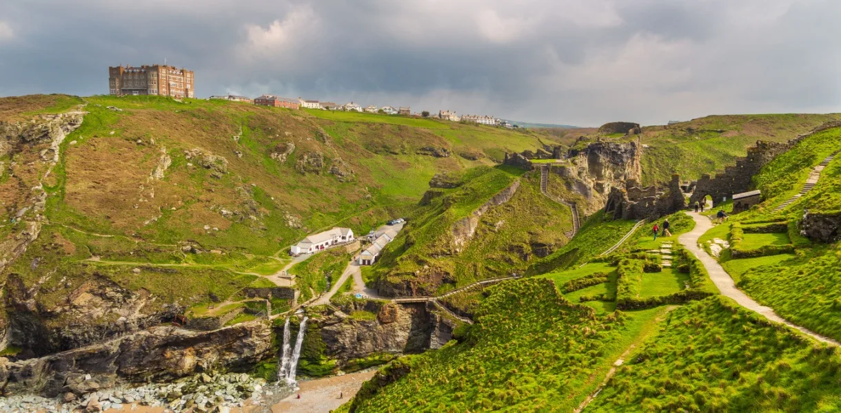 Wide view of Tintagel Castle in Cornwall with ruined stone walls and winding paths spread across steep green cliffs above a rocky cove under dark cloudy skies