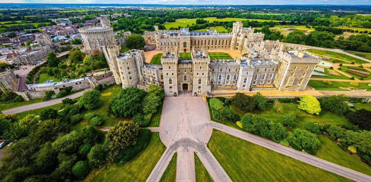 Aerial view of Windsor Castle with its stone towers, battlements and large central courtyard, surrounded by formal gardens, tree-lined grounds and the town and green fields beyond.
