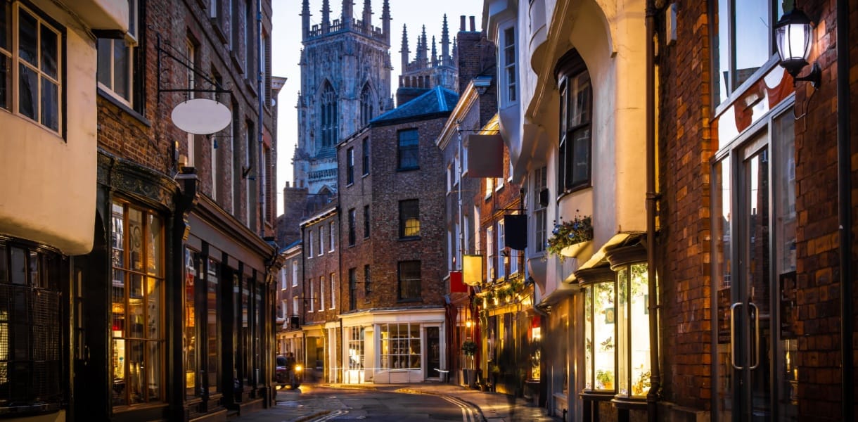 A narrow cobbled street in York at dusk, lined with historic brick and timber-fronted buildings, with warm shop lights glowing and York Minster rising in the background.