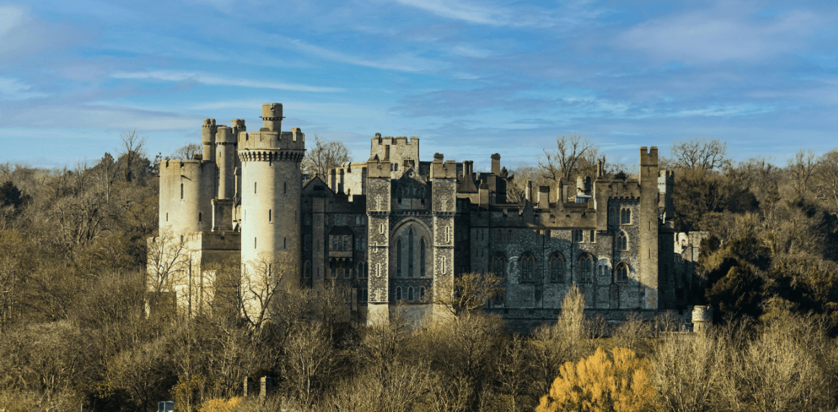 Arundel Castle in West Sussex rising above surrounding trees, with its towers, battlements and historic stone façade set against a blue sky.