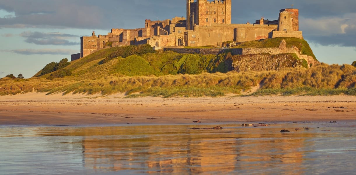Bamburgh Castle glowing in warm late-day light above sandy dunes and a wide beach, with its stone walls and towers reflected in the wet sand below.