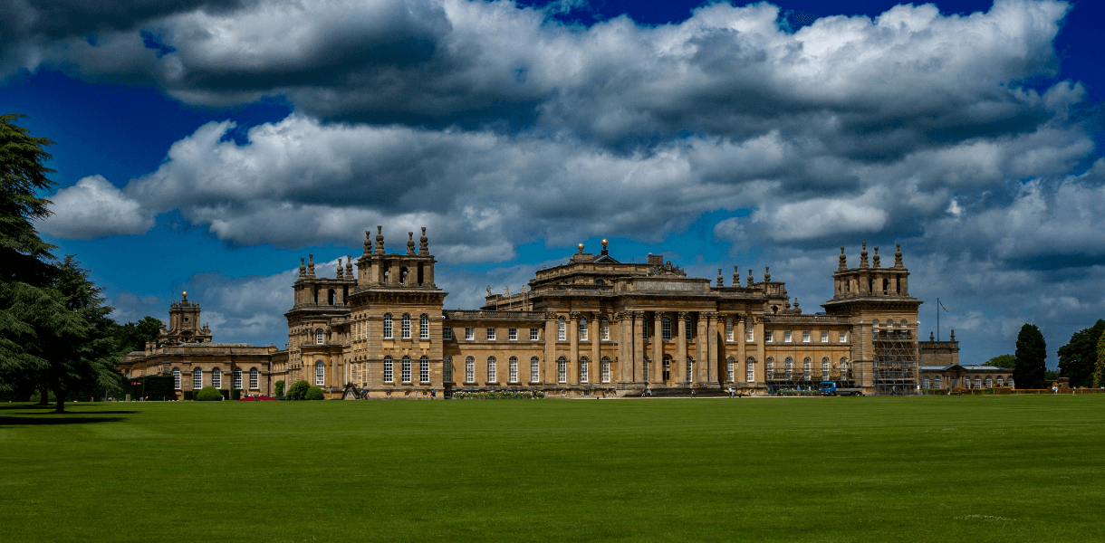 Wide view of Blenheim Palace in Oxfordshire, with its grand honey-coloured Baroque façade set behind an open green lawn beneath dramatic billowing clouds.