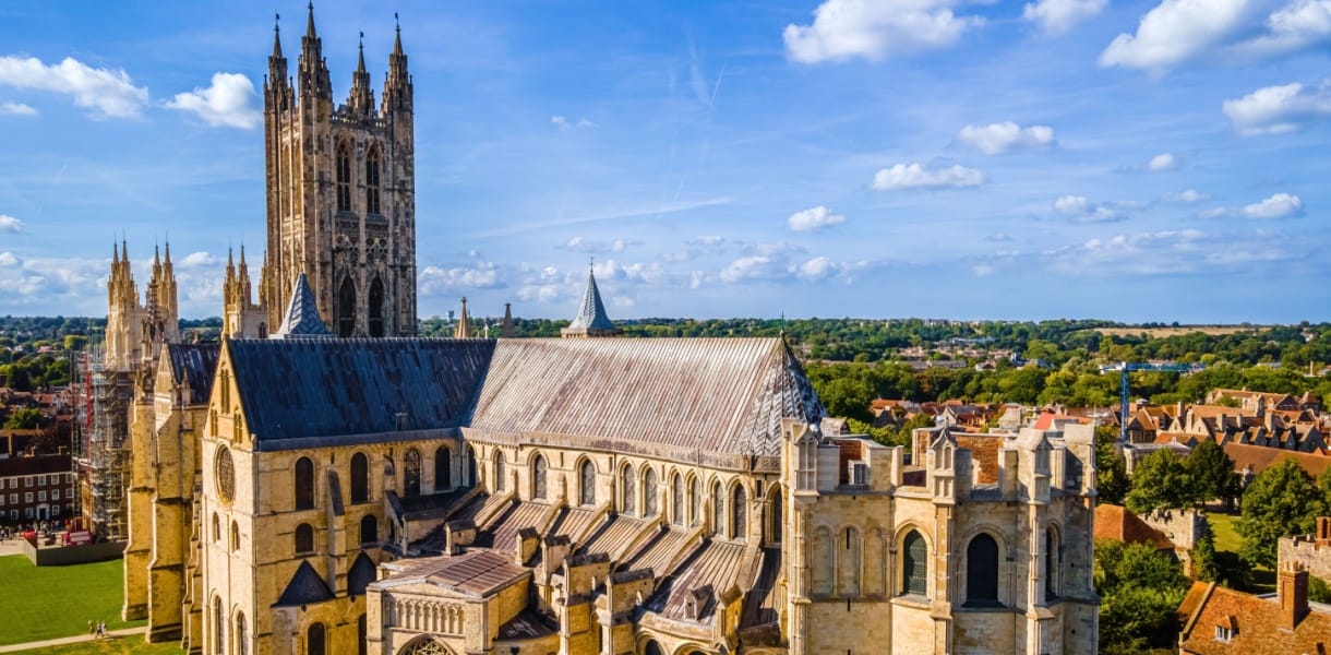 Canterbury Cathedral rising above the rooftops of Canterbury, with its Gothic towers, historic stone nave and surrounding Kent countryside under a blue sky.