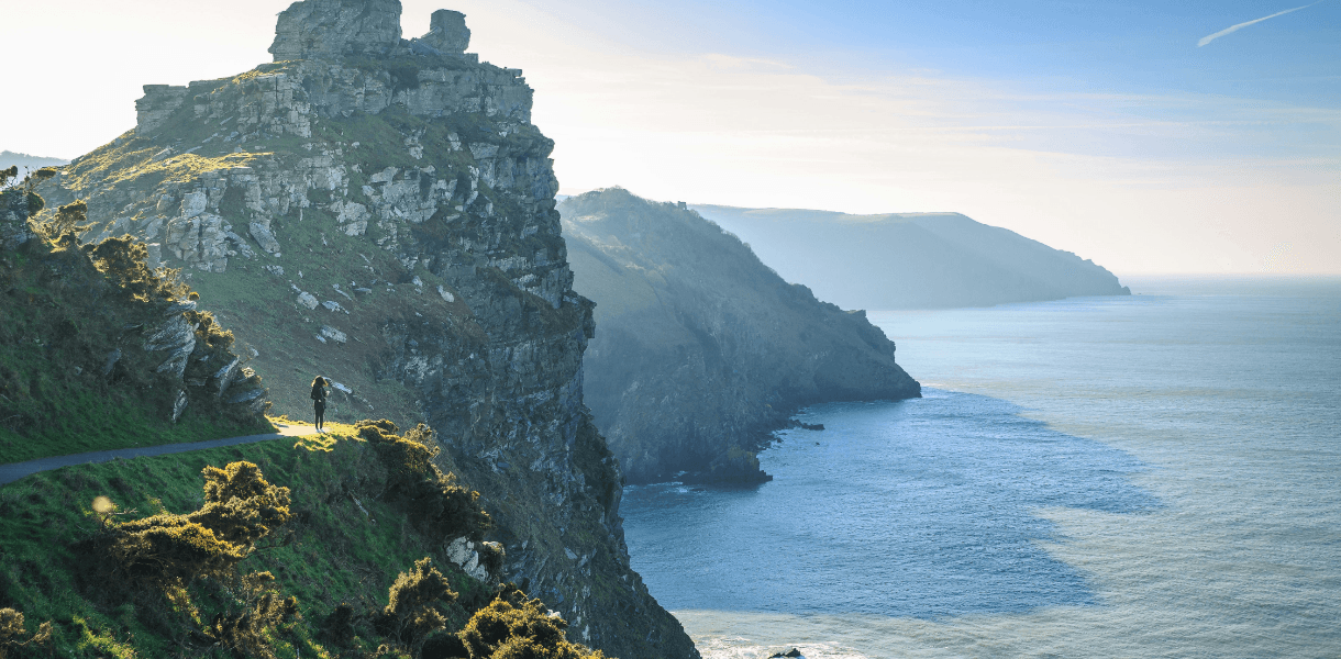 Rugged Devon coastline with steep green cliffs, rocky headlands and a lone walker on a coastal path overlooking the sea.