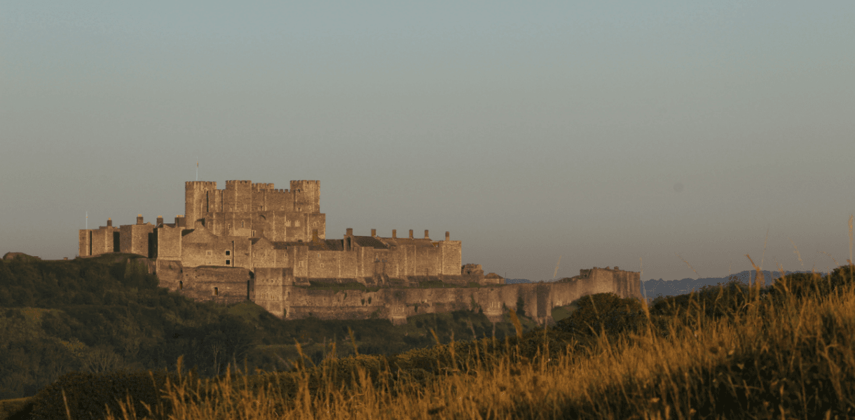 Dover Castle standing on a hill above the Kent countryside, with its medieval walls and keep lit by warm evening light.