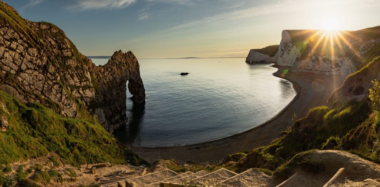 Durdle Door sea arch on the Jurassic Coast in Dorset, with chalk cliffs, a sheltered beach and golden sunlight over the sea.