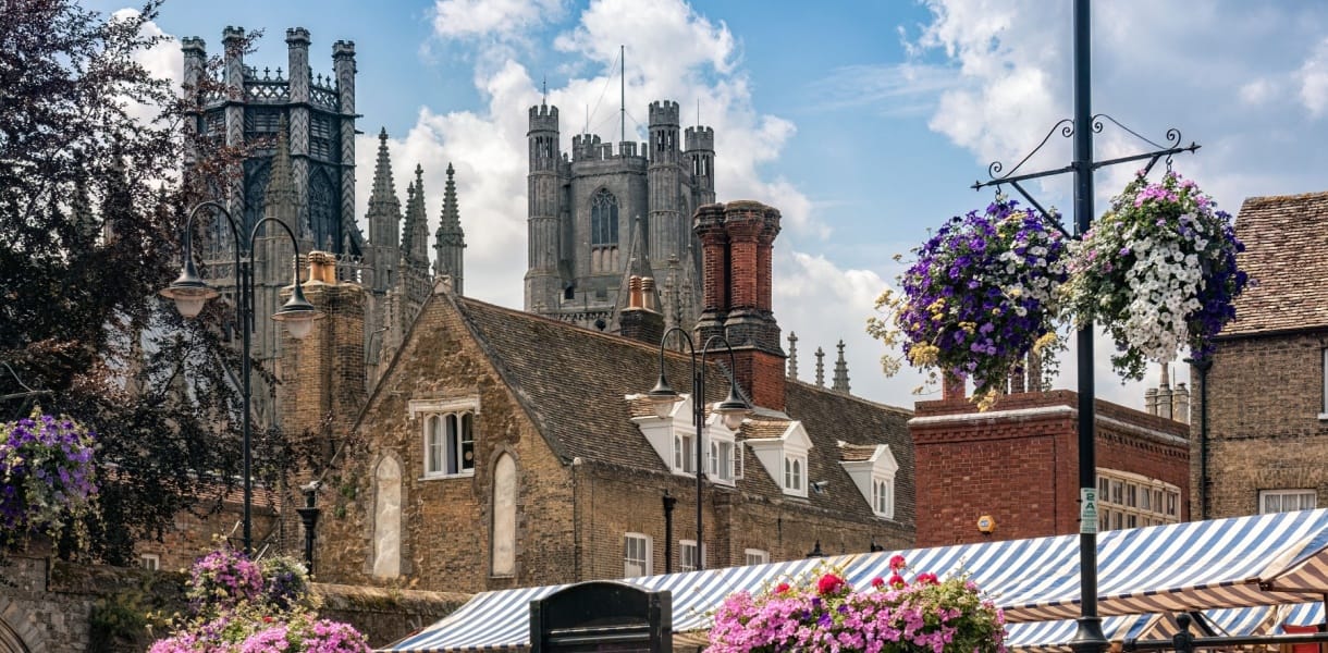 View of Ely city centre with historic stone buildings, flower baskets and market stalls in the foreground, with the towers of Ely Cathedral rising above the rooftops under a bright sky.