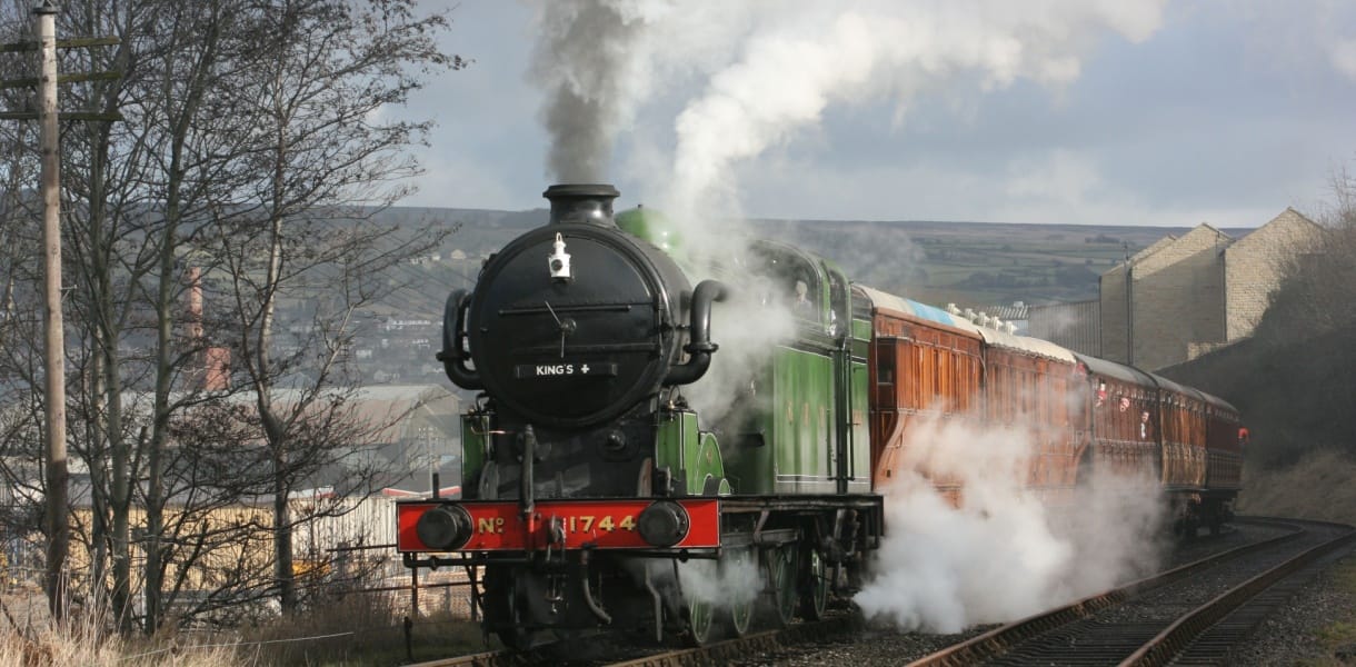 Green steam locomotive pulling vintage carriages along a heritage railway line, with clouds of steam rising against a countryside backdrop.