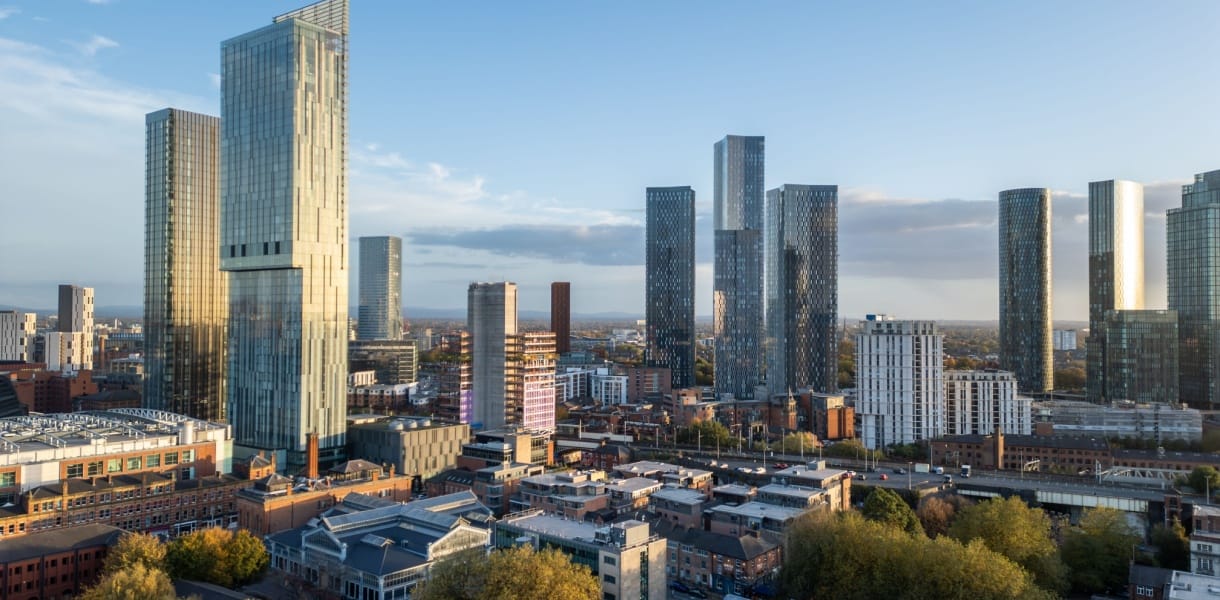 A wide skyline view of central Manchester in clear daylight, showing a cluster of tall glass skyscrapers rising above lower red-brick buildings, roads, and patches of autumn trees under a pale blue sky.