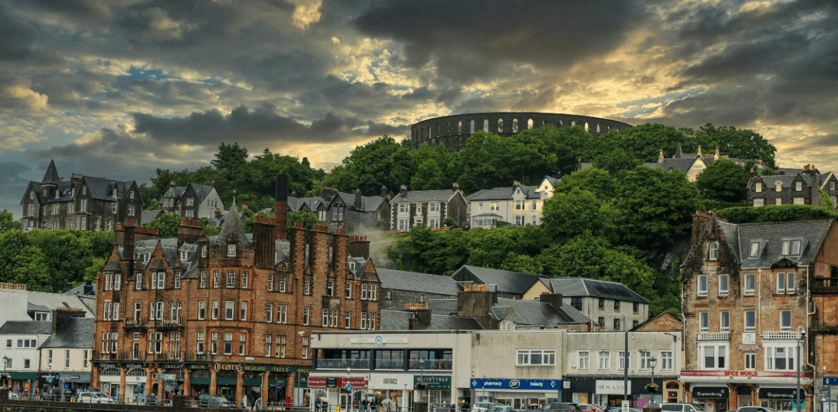 Oban town centre in Scotland with Victorian buildings, wooded hillside homes and McCaig’s Tower overlooking the town beneath a dramatic cloudy sky.