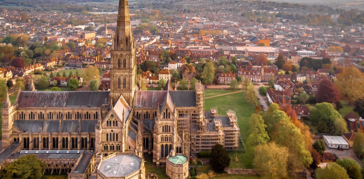 Aerial view of Salisbury Cathedral and its tall spire, with the historic city, green spaces and surrounding countryside beyond.