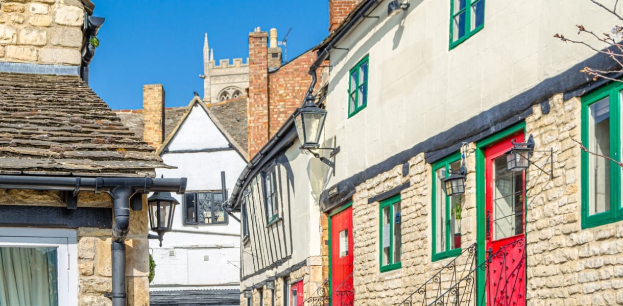 Sunlit stone cottages and narrow old buildings in Stamford, with bright red and green painted doors and windows, and a church tower rising in the background against a clear blue sky.