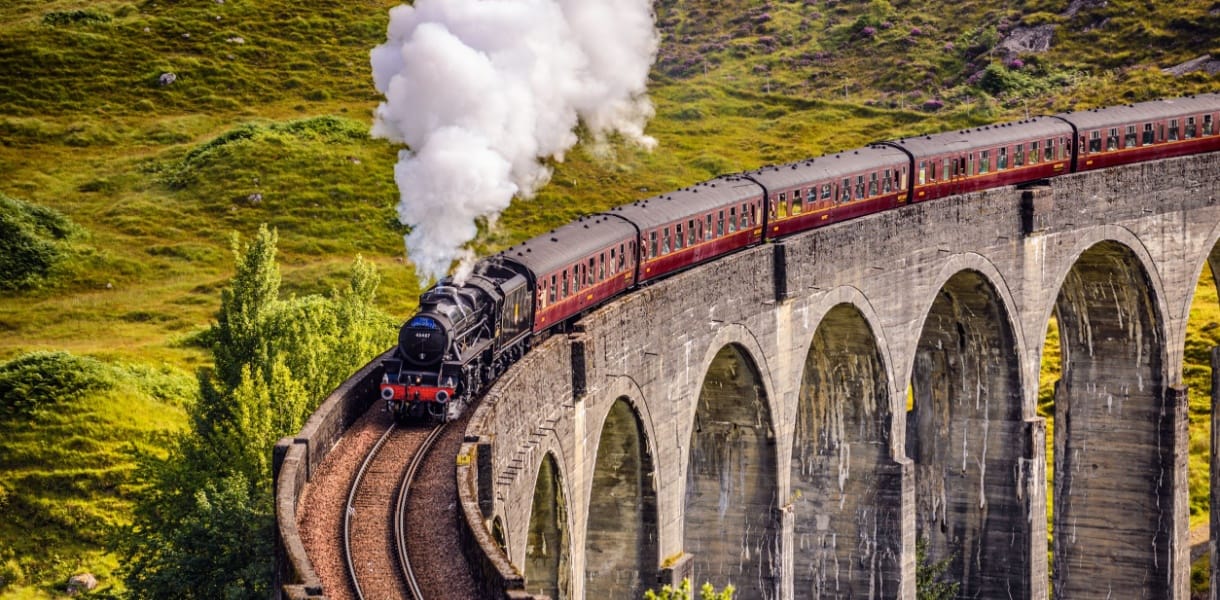 Steam train crossing the Glenfinnan Viaduct in the Scottish Highlands, with white billowing smoke, maroon carriages and green hills surrounding the sweeping railway bridge.