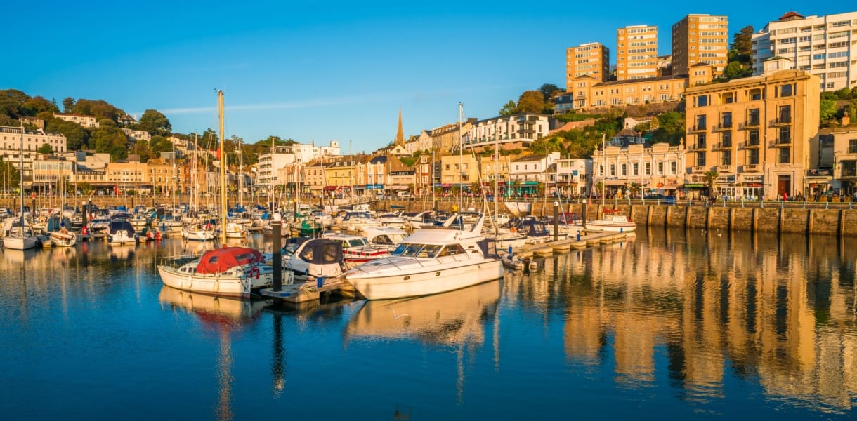 Boats moored in Torquay harbour at golden hour, with waterfront buildings and hillside homes reflected in the calm water.