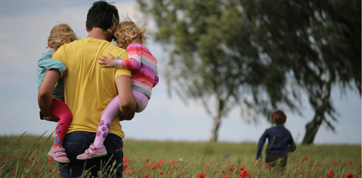 Adult carrying two small children through a grassy field dotted with red wildflowers, while another child walks ahead towards trees under a cloudy sky.