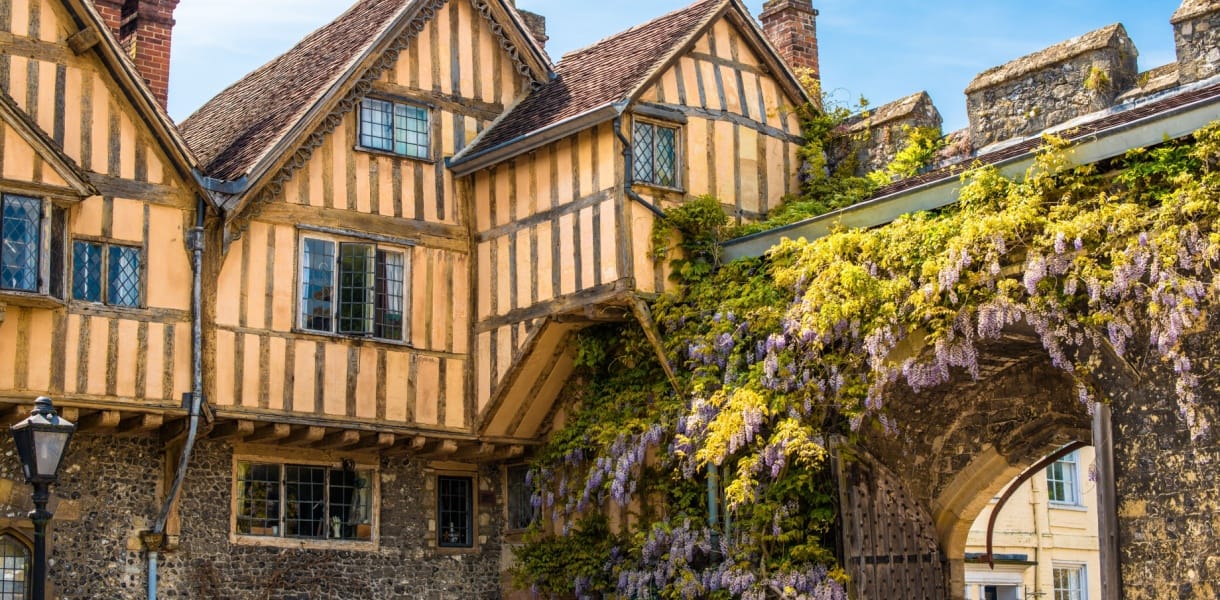 Historic timber-framed buildings in Winchester beside a stone archway covered with flowering wisteria.