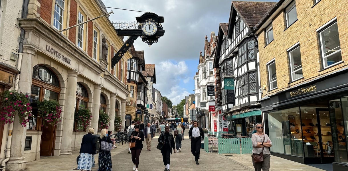 Pedestrian shopping street in Winchester with historic brick and timber-framed buildings, an ornate hanging clock and people walking past shops and cafés.