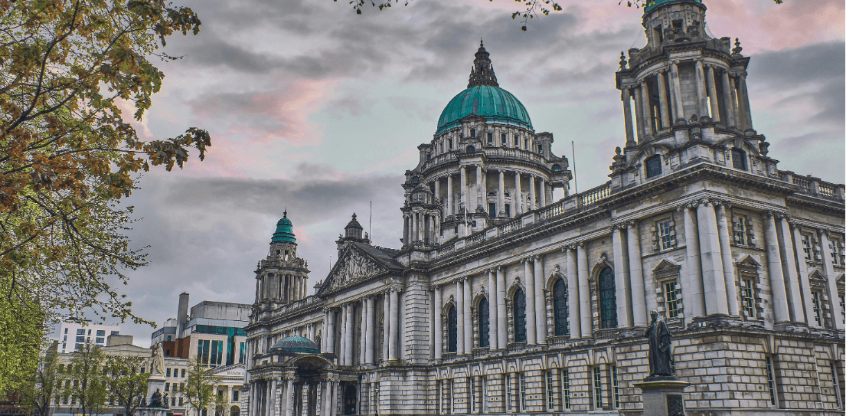 Belfast City Hall with its green copper dome and ornate stone façade, seen from the surrounding lawns beneath a cloudy evening sky and framed by leafy trees.