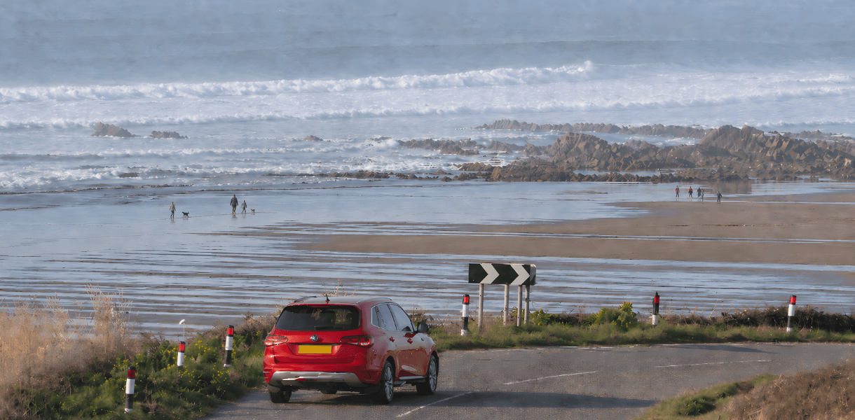 Red car driving along a winding coastal road above a wide sandy beach, with waves, rocky outcrops, distant walkers and dog walkers on the shoreline.