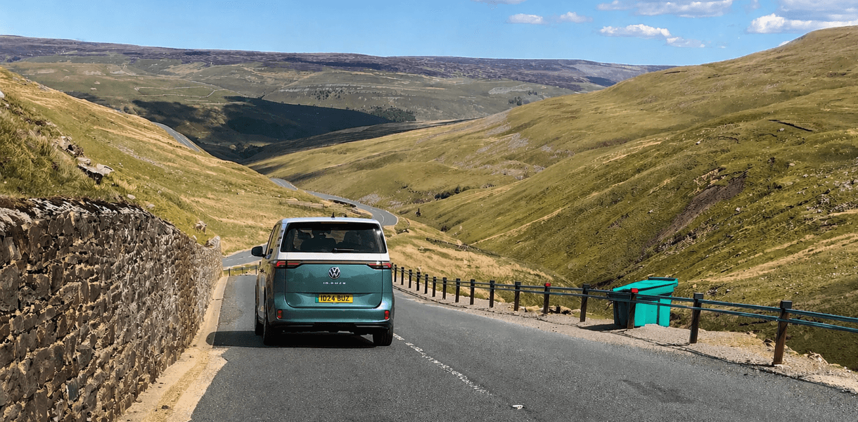 VW ID Buzz driving down a winding road through the rolling hills and open moorland of the Yorkshire Dales on a bright sunny day.