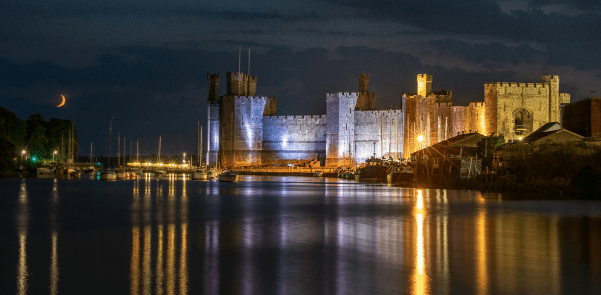 Caernarfon Castle illuminated at night beside the waterfront, with boats, reflections on the water and a crescent moon in the dark sky.