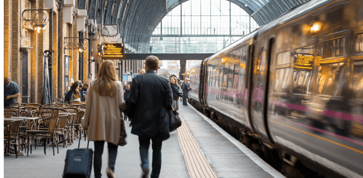 Two travellers walk along a busy railway platform with luggage beside a train, beneath the arched glass roof of a large station with café seating and departure boards in view.