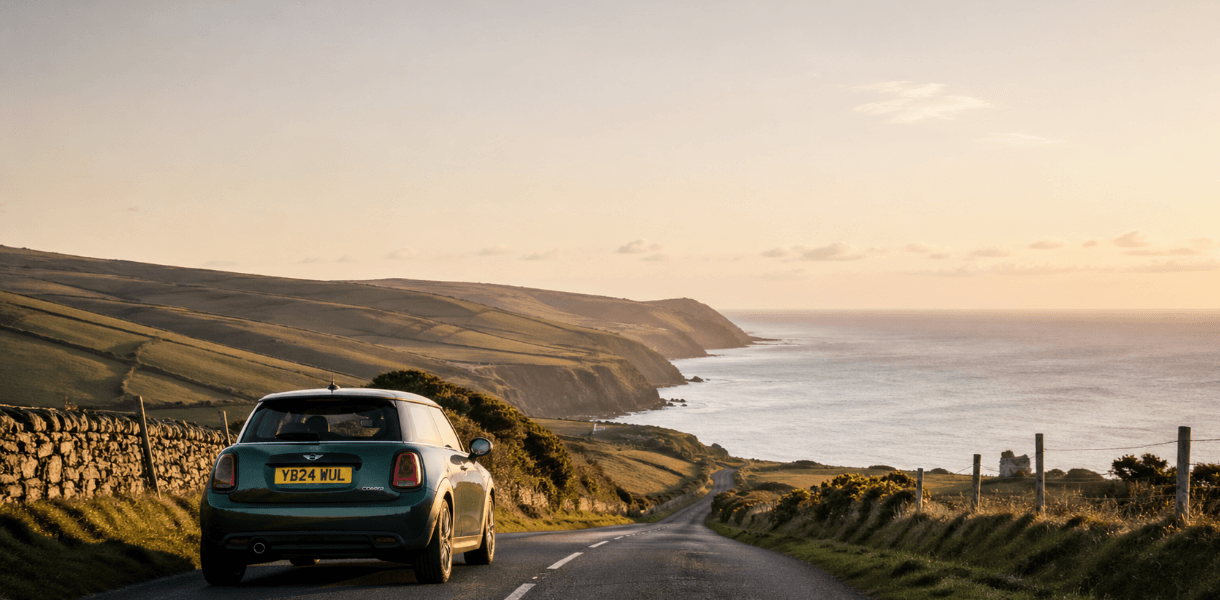 A green mini car on a quiet coastal road at golden hour, with rolling British countryside, stone walls and cliffs leading down to the sea beneath a wide open sky.