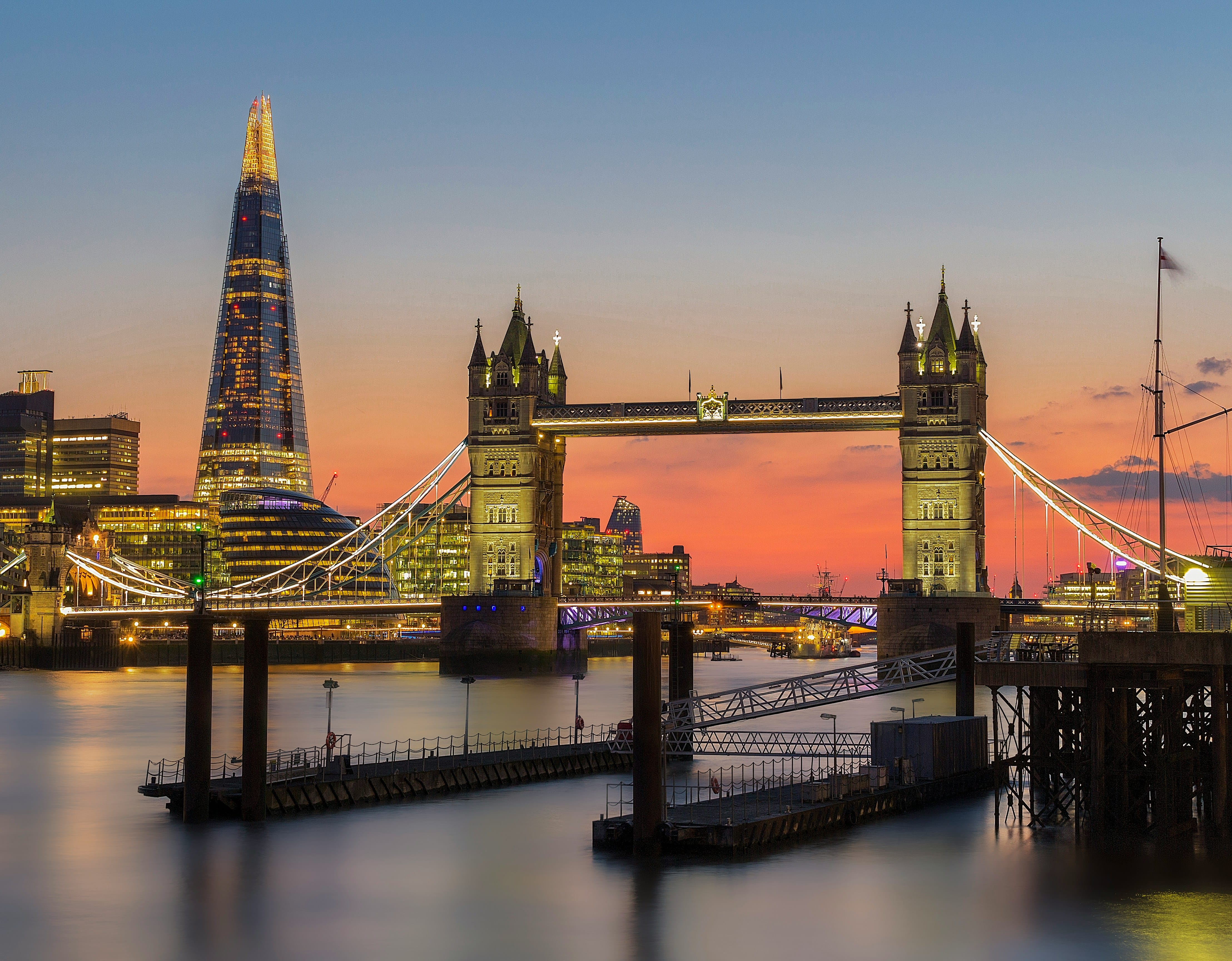 Tower Bridge and The Shard lit up at dusk in London, seen across the River Thames, with calm water in the foreground and a soft orange and blue evening sky.